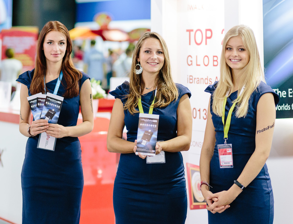 Drei Hostessen in blauen Kleidern stehen auf einer Messe und halten Informationsflyer in den Händen.