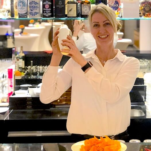 Barkeeperin in weißem Hemd steht hinter der Bar und mixt einen Cocktail mit einem Shaker